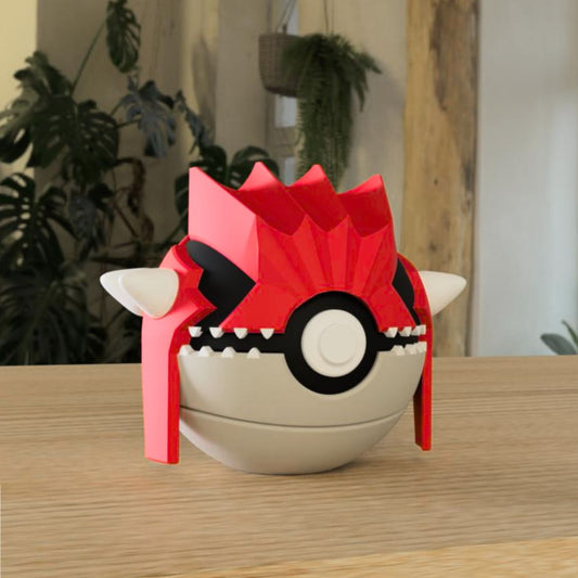 3D printed Groudon-themed Pokéball replica on a wooden surface, with green plants and a window in the blurred background. The Pokéball features Groudon's signature red and black patterns, with white accents.
